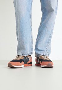 Zapatillas deportivas con una mezcla de materiales negros, naranjas y beiges. Presentan una parte superior texturizada, suela de goma y cordones beiges. Diseño de punta redondeada.