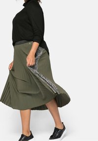 Jupe plissée vert olive avec une taille noire et un accent latéral de motifs noir et blanc. Associée à un haut noir et des baskets.