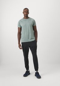 Grünes meliertes Sport-T-Shirt mit kurzen Ärmeln, kombiniert mit schwarzen taillierten Jogginghosen und blauen Sportschuhen auf einem schlichten Hintergrund.