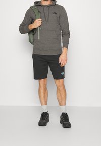 Felpa grigia con logo, pantaloni corti neri con logo, calzini alla caviglia grigi e scarpe sportive nere. Zaino in materiale verde.