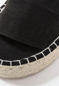 Sandale en suede noir avec une large sangle et une semelle en jute enroulée avec des coutures blanches. Design plat avec une texture lisse.