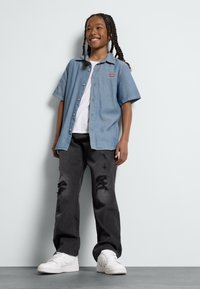 Lichtblauw overhemd met korte mouwen, witte t-shirt met ronde hals, zwart versleten jeans en witte sneakers gedragen door een kind.