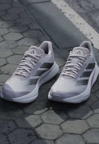 Paar graue Adidas Laufschuhe mit schwarzen Streifen auf sechseckigem Pflaster nahe einem Straßenbordstein.