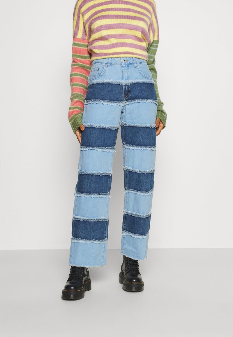 Vaqueros azules de mezclilla con rayas horizontales de tonos variados y bordes sin rematar. Combinados con un suéter multicolor de rayas y botas negras.