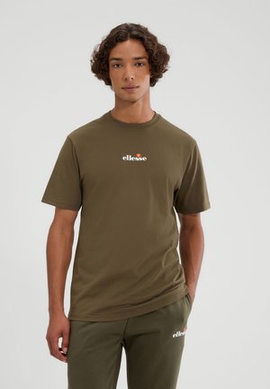 Joven con cabello rizado que lleva puesta una camiseta y pantalones ellesse verde oliva a juego, de pie contra un fondo liso y claro.