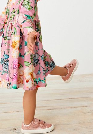 Robe rose à motif floral avec manches longues, associée à des baskets roses pailletées avec double bride Velcro. Fond à texture bois.
