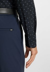 Langarm zwart shirt met blauw ruitpatroon, met knoopmanchetten, gecombineerd met marineblauwe broek met een zwarte riem en een klein merklabel.