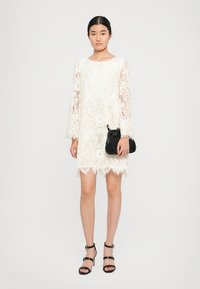 Weißes Spitzenkleid mit langen Ärmeln, durchscheinenden Spitzenakzenten, gewelltem Saum, kombiniert mit einer schwarzen strukturierten Handtasche und schwarzen Riemchensandalen.