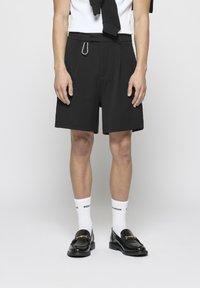 Shorts noirs ajustés avec une texture lisse, dotés de plis sur le devant, de passants pour ceinture et d'un clip en métal. Associés à des chaussettes blanches et des mocassins noirs.