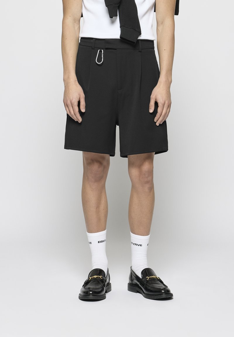 Shorts noirs ajustés avec une texture lisse, dotés de plis sur le devant, de passants pour ceinture et d'un clip en métal. Associés à des chaussettes blanches et des mocassins noirs.