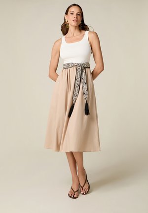 Vrouw draagt een mouwloos wit topje en een beige midirok met een zwart-witte patroonriem en bruine sandalen, staand tegen een effen achtergrond.