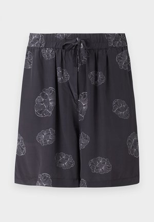 Νέος άνδρας με μαύρο κοντομάνικο πουκάμισο με λουλούδια και ταιριαστό σορτς πάνω από λευκή μπλούζα, λευκά παπούτσια slip-on, στέκεται με το ένα χέρι στην τσέπη.