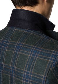 Wełniana kurtka z ciemnoniebieskim kołnierzem, w zielono-niebieską kratę. Faktura wydaje się miękka i ciepła, z widocznymi szczegółami przeszyć.