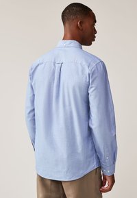 Hellblaues langärmliges Hemd aus weichem Stoff, mit Knopfkragen, einfacher Rückenpasse und geknöpften Manschetten.