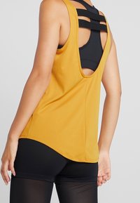 Canotta sportiva gialla con scollatura arrotondata, abbinata a leggins neri. La canotta presenta una superficie liscia e un orlo curvo.