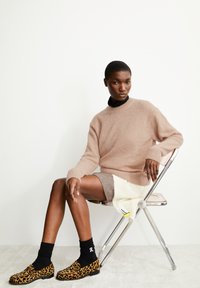 Beige gebreide trui met geribde manchetten, lichtgrijze rok, zwarte sokken en schoenen met luipaardprint. Zittend op een chromen stoel, minimalistische achtergrond.