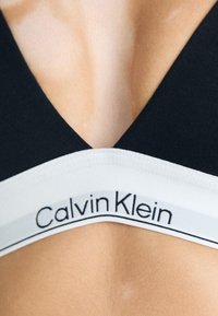 Zwarte bralette met een V-halsontwerp. Voorzien van een grijze en witte elastische band met het 'Calvin Klein' logo. Zachte, gladde textuur.
