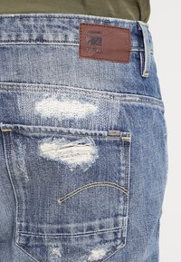 Vue arrière de jeans en denim bleu avec des patchs déchirés, une poche arrière et une étiquette de marque G-Star RAW en cuir marron sur la ceinture.