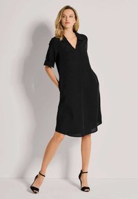 Schwarzes Kurzarmkleid mit Kragen, Seitentaschen und lockerem Schnitt. Hergestellt aus einem leichten Stoff mit glatter Textur.