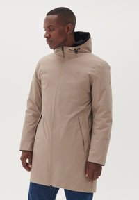 Abrigo impermeable beige con cierre frontal, dos bolsillos laterales, capucha ajustable y textura suave, acompañado de pantalones de mezclilla azul.
