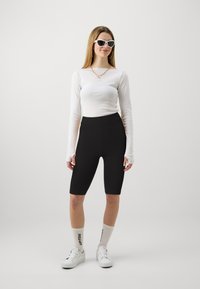 Biely dlhý rukáv, čierne cyklistické šortky s vysokým pásom, biele tenisky a podkolienky s textom; model má na sebe biele slnečné okuliare.