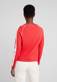 Felpa rossa con strisce bianche sui lati, colletto a girocollo e maniche raglan. Texture morbida e design aderente, abbinata a pantaloni chiari.