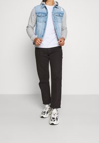 Chaleco de mezclilla con capucha gris, de lavado claro, con dos bolsillos en el pecho; camiseta blanca lisa, pantalones negros y zapatillas negras/blancas con acentos estampados.