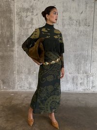 Zelený pletený top s fringingom, kombinovaný s vzorovanou priliehajúcou sukňou. Doplnky zahŕňajú hnedú semišovú kabelku a béžové lodičky so cvočkami.