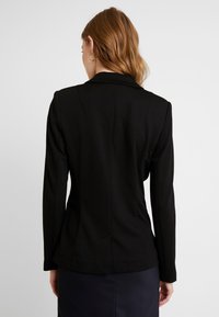 Svart skräddarsydd kavaj med en åtsittande design, långa ärmar och klippt krage. Slät tyg, detaljerad sömnad och en elegant silhuett.