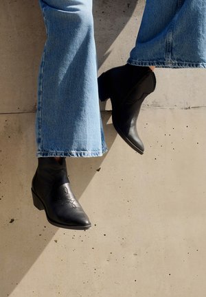 Czarne skórzane botki za kostkę z szpiczastymi noskami, ozdobnym przeszyciem i słupkowym obcasem, w zestawie z niebieskimi spodniami typu wide-leg.