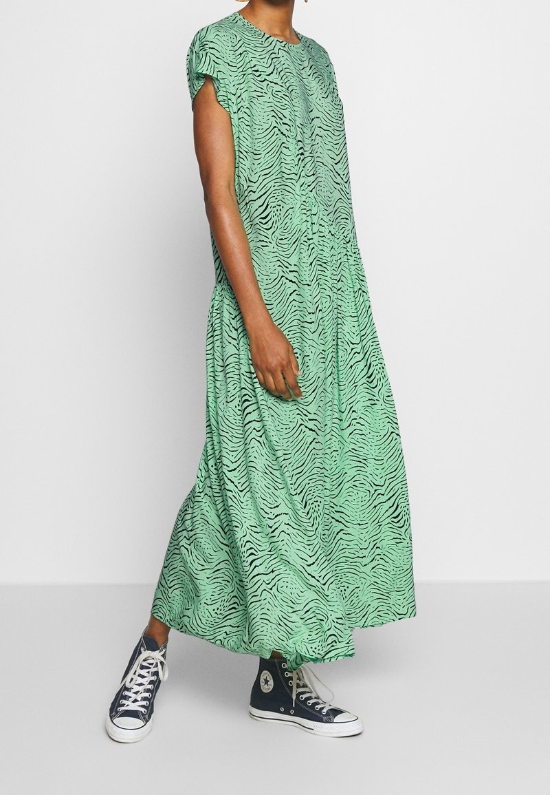 Personne portant une robe verte à manches courtes, longue jusqu'aux chevilles, avec des rayures zébrées noires et des baskets montantes noires avec des lacets blancs.