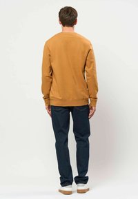 Homme portant un sweat-shirt marron à manches longues, un jean bleu foncé et des chaussures blanches, debout face à un fond blanc uni.