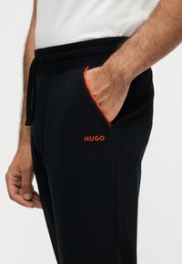 Gros plan d'une personne portant un pantalon de survêtement noir avec un logo orange "HUGO" et la main dans la poche avant.