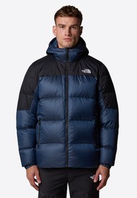 Granatowa kurtka puchowa z czarnymi akcentami, zasuwana z przodu, z kapturem. Charakteryzuje się teksturowanym, izolowanym designem oraz bocznymi kieszeniami.