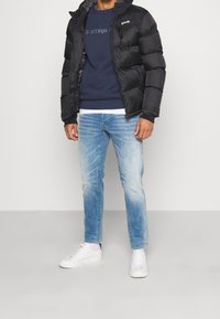 Man som bär en svart puffjacka, marinblå sweatshirt, ljusblå jeans och vita sneakers mot en enfärgad grå bakgrund.