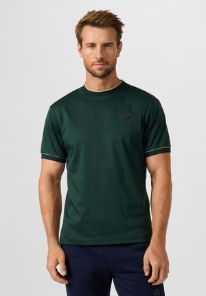 Man met kort bruin haar draagt een donkergroen T-shirt met korte mouwen en een marineblauwe broek, staand tegen een effen lichte achtergrond.
