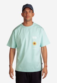 Minttu vihreä puuvillapaita lyhyillä hihoilla, jossa on etupuolella teksti "KOHTI AURINKOA" ja keltainen auringonkukkagrafiikka.