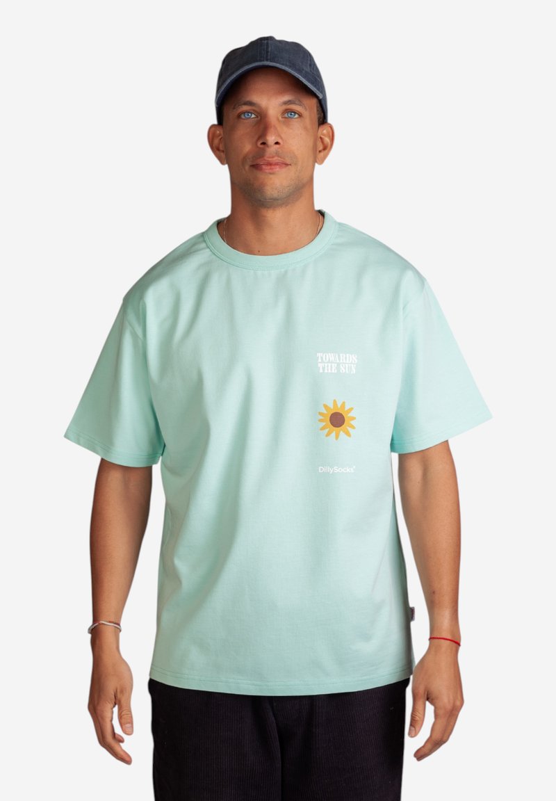 Minttu vihreä puuvillapaita lyhyillä hihoilla, jossa on etupuolella teksti "KOHTI AURINKOA" ja keltainen auringonkukkagrafiikka.