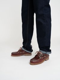 Des chaussures de bateau en cuir brun avec un bout arrondi, des coutures blanches et des détails en lacets, associées à un jean en denim foncé retroussé aux chevilles.