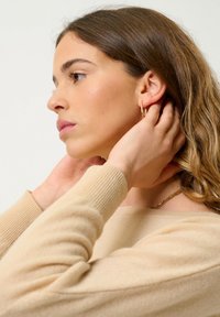 Maglione beige a maglia con polsini a costine, orecchini a cerchio dorati e una delicata collana a catena. Capelli in morbide onde, mostrati di profilo.