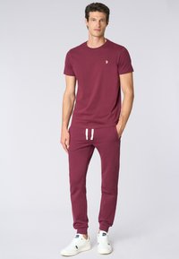 T-shirt color bordeaux e pantaloni sportivi abbinati in cotone, con un piccolo logo sul petto e polsini elastici. Abbinati a sneaker bianche.