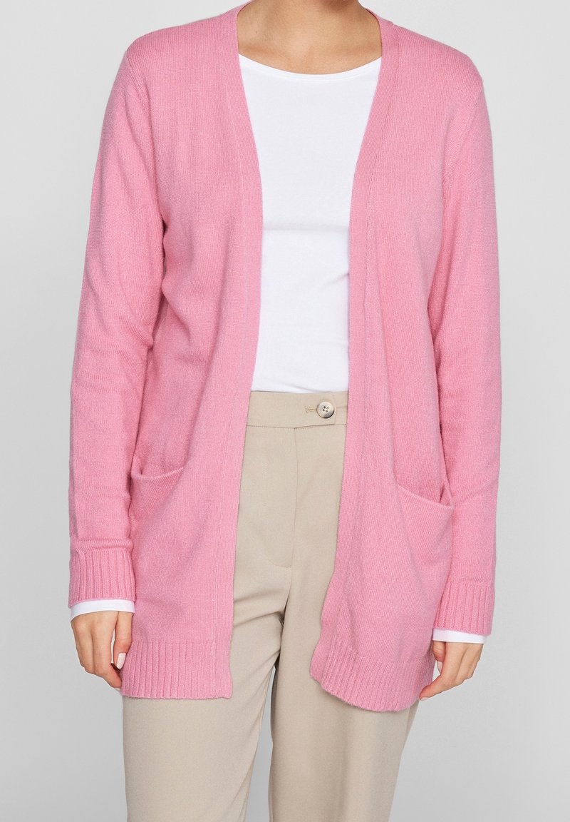 Femme portant un cardigan rose clair ouvert sur le devant avec des poches à l'avant, par-dessus une chemise blanche et un pantalon beige.