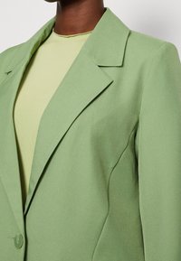 Blazer verde dal design strutturato, con rever a incavo, chiusura con un solo bottone e texture liscia, abbinato a una maglietta verde chiaro sotto.
