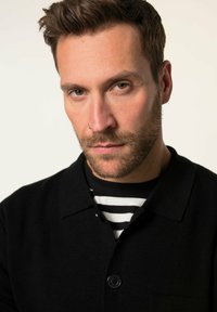 Camicia nera con colletto, indossata sopra una maglietta a righe bianche e nere. La camicia presenta un tessuto testurizzato e una tasca sul petto.