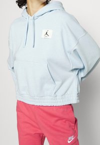 Ljusblå kort hoodie med en känguruficka, ribbad fåll och Jordan-logotyp på bröstet; bärs med rosa svettbyxor.