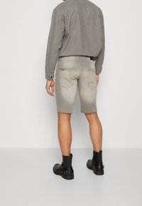 Shorts in denim grigio con una finitura sbiadita e orli arrotolati, abbinati a stivaletti neri alla caviglia. Il modello indossa una camicia grigia con bottoni infilata dentro.