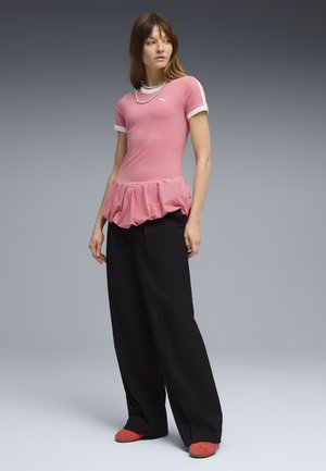 Jonge vrouw met een roze top met gerimpelde zoom, zwarte wijde broekspijpen, rode schoenen en een parelketting tegen een grijze verloop achtergrond.