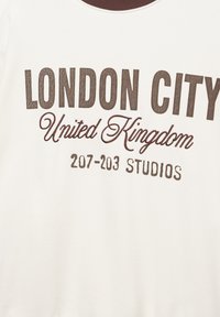 Sweatshirt blanc avec texte marron. Le devant présente « LONDON CITY » et « United Kingdom » en polices gras, avec « 207-203 STUDIOS » en dessous.