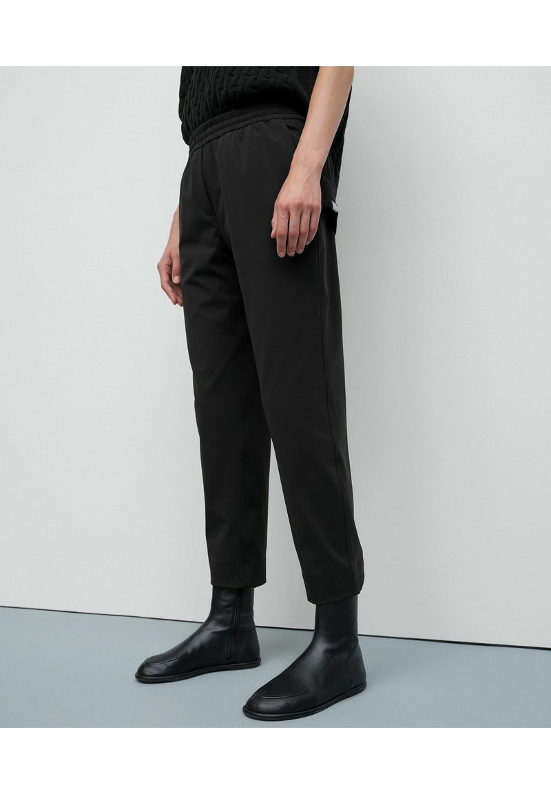 Person trägt schwarze, gekürzte Hosen und schwarze Leder-Stiefeletten, steht vor einer hellgrauen Wand und einem hellgrauen Boden.