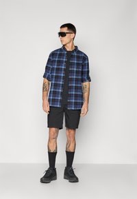 Plaid shirt in blue and black, short sleeves, paired with black cargo shorts. Black socks and shoes complete the look. Sleek sunglasses worn.